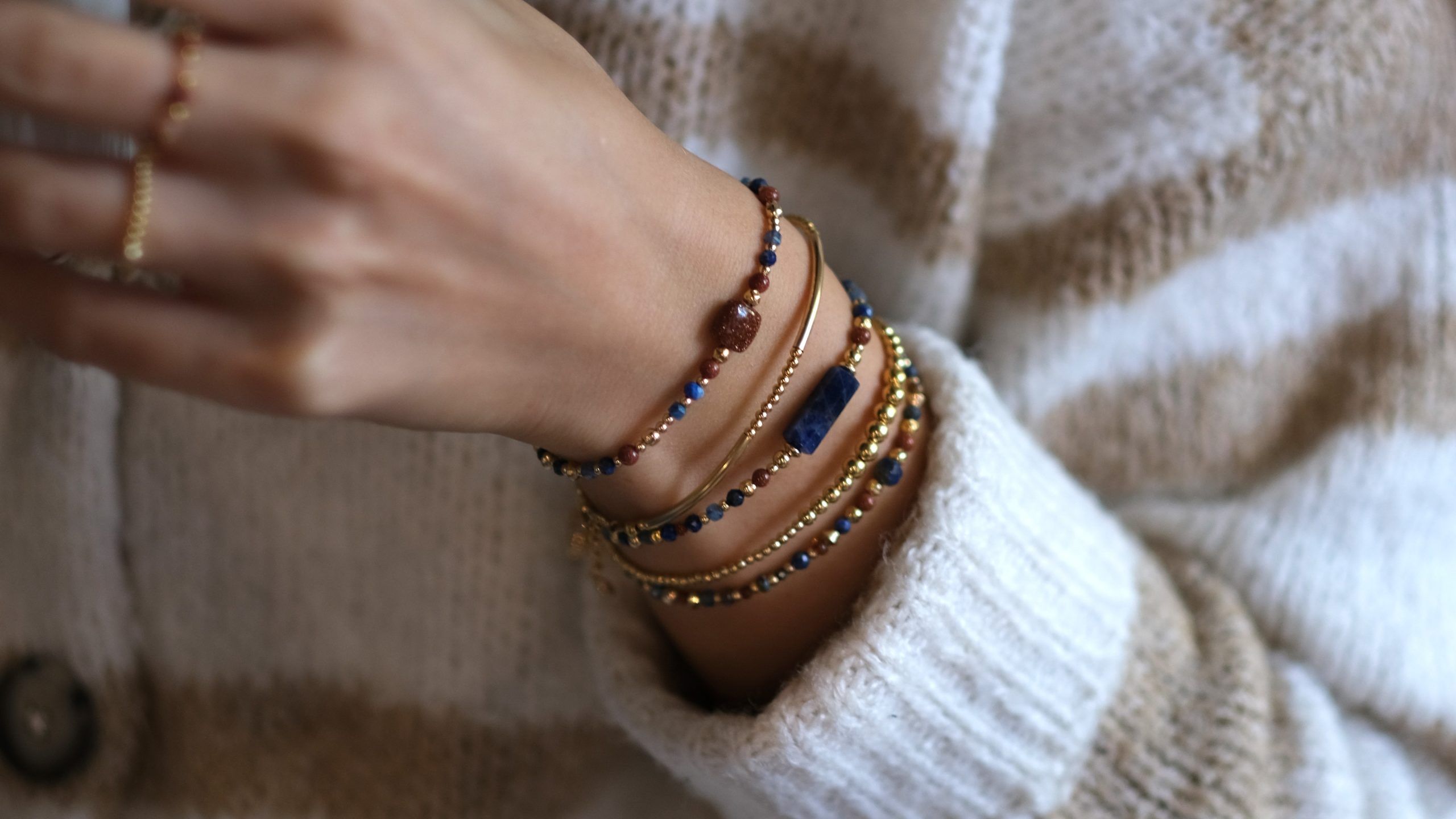 Poignet paré de bracelets en or laminé, perles de lapis-lazuli et pierre de soleil, sur un pull rayé beige